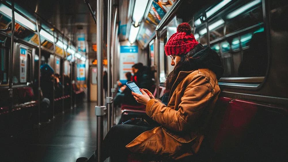 A young woman wearing a red knit hat and a brown coat sits alone in a subway car, engrossed in her smartphone. The subway car is dimly lit with a few other passengers scattered around, creating a cozy and quiet atmosphere. The woman appears focused and comfortable as she navigates her mobile device.