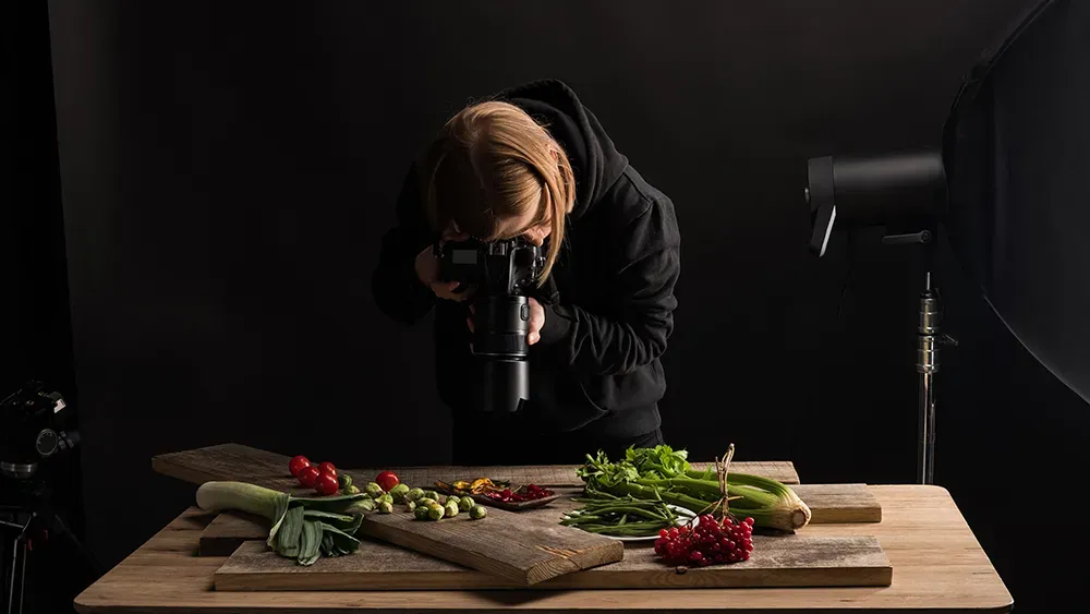 A photographer wearing a black hoodie is leaning over a table with a camera, taking close-up shots of fresh vegetables and fruits arranged on wooden boards. The scene is set in a studio with professional lighting equipment.