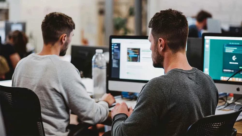 Two men working on desktop computers in a modern office setting. They are focused on their screens, one of which displays code and the other showing a user interface. The workspace has a collaborative environment with open desks and minimal partitions.