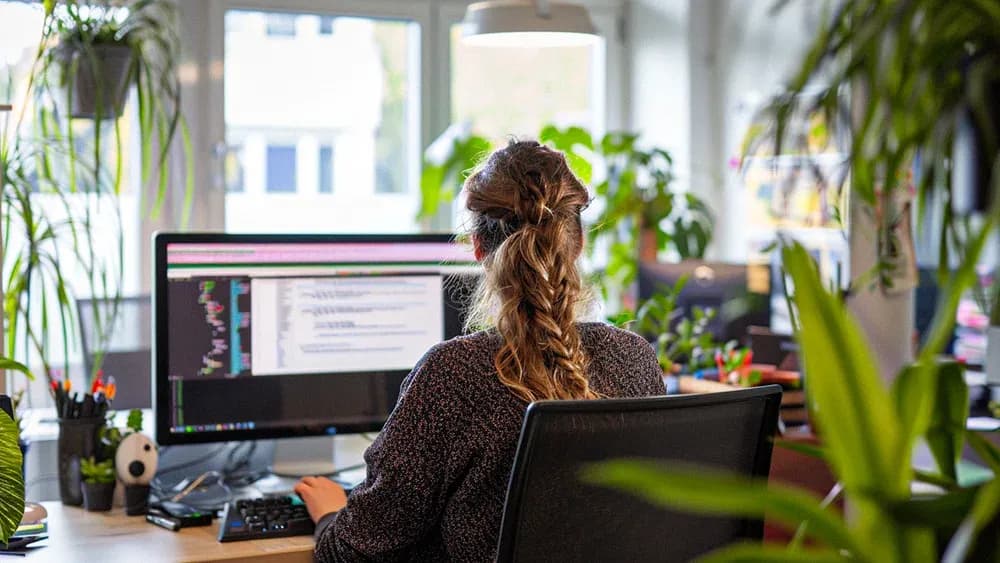 A woman with long hair, tied in a braid, working on a computer in a plant-filled office. She is focused on coding displayed on a large monitor, with various office supplies and green plants surrounding her workspace.