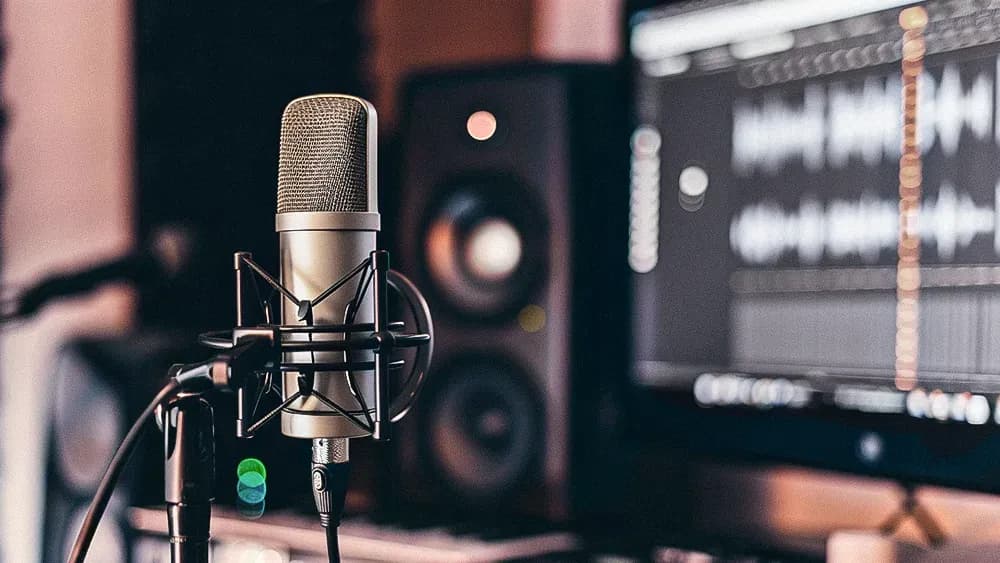 A professional condenser microphone set up in front of a computer screen displaying audio waveforms. The background includes studio speakers, suggesting a recording or podcasting environment.