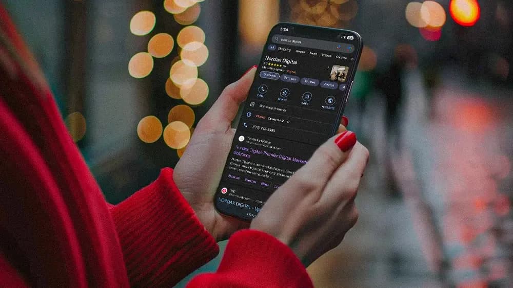 A person wearing a red sweater holds a smartphone, searching for "Nordax Digital" on a search engine. The phone screen displays search results with Nordax Digital listed. The background is blurred, with bokeh lights creating a festive and cozy atmosphere, suggesting an evening or night setting.