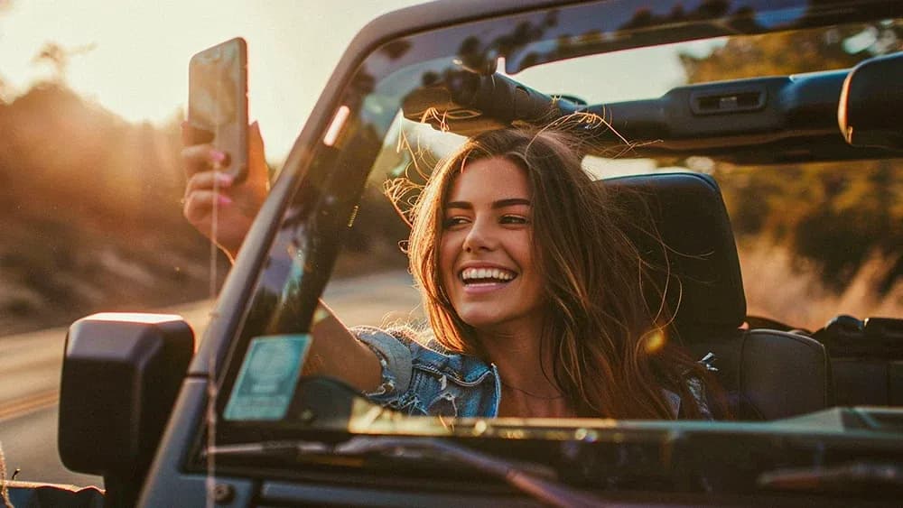 A young woman with long hair and a bright smile takes a selfie while driving in a convertible with the top down. The sun is setting, casting a warm golden glow over the scene. The woman looks carefree and happy, enjoying the open road and capturing the moment on her smartphone.