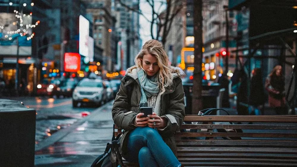 A young woman with blonde hair is sitting on a bench in a bustling city street, intently looking at her smartphone. She is dressed in a winter coat and scarf, and the street is adorned with lights and moving cars, capturing the essence of urban life.
