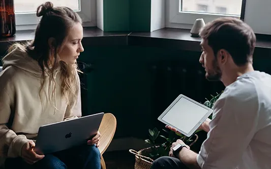 A young woman and a man are engaged in a discussion while holding digital devices. The woman, wearing a beige hoodie, holds a MacBook, while the man, in a white shirt, holds an iPad. They are seated near a window, in a cozy, modern setting with some plants and decor visible.