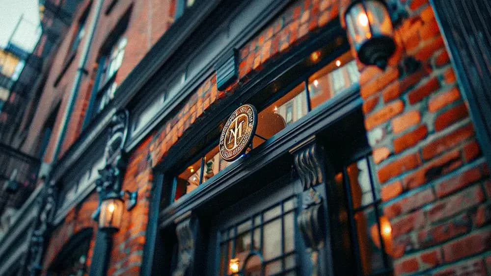 A close-up view of the entrance to a building with a sign that reads "M" above the door. The building features red brick walls and black-framed windows with warm lighting visible inside.