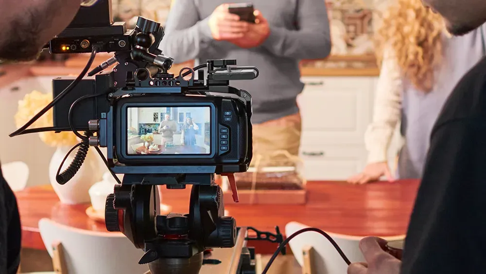 A professional video camera is set up in a cozy kitchen environment, capturing footage of people engaged in a conversation. The camera's screen shows the scene being recorded, with a focus on the individuals in the background.